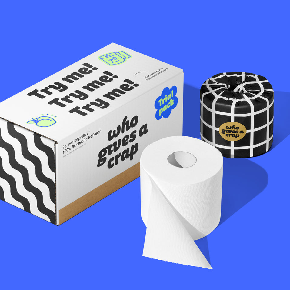 On a bold blue background sits one wrapped roll and one unwrapped roll of bamboo Who Gives a Crap toilet paper next to a trial pack box with Try me printed three times.