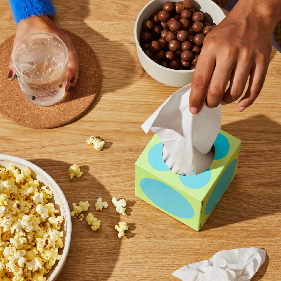 A hand reaches for a tissue from a blue and green box of tissues on a table with food and drinks.