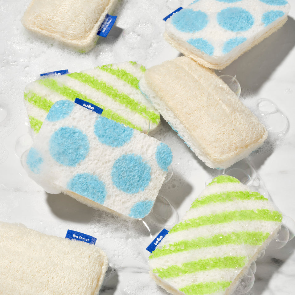 Multiple Who Gives A Crap scrub sponges scattered in soapy water, displaying the blue polka dot, green stripe and natural loofah sides