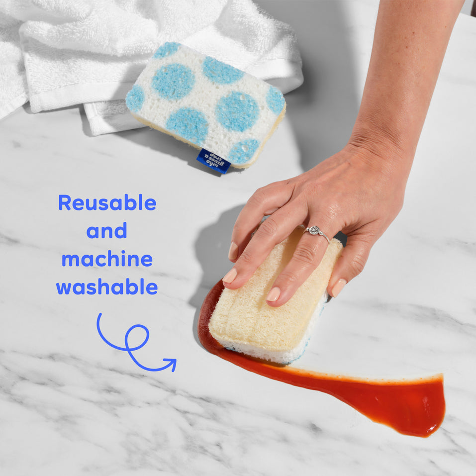 A hand scrubbing a surface with a Who Gives A Crap natural loofah scourer sponge next to a blue polka dot sponge, with a callout highlighting that the sponges are reusable and machine washable