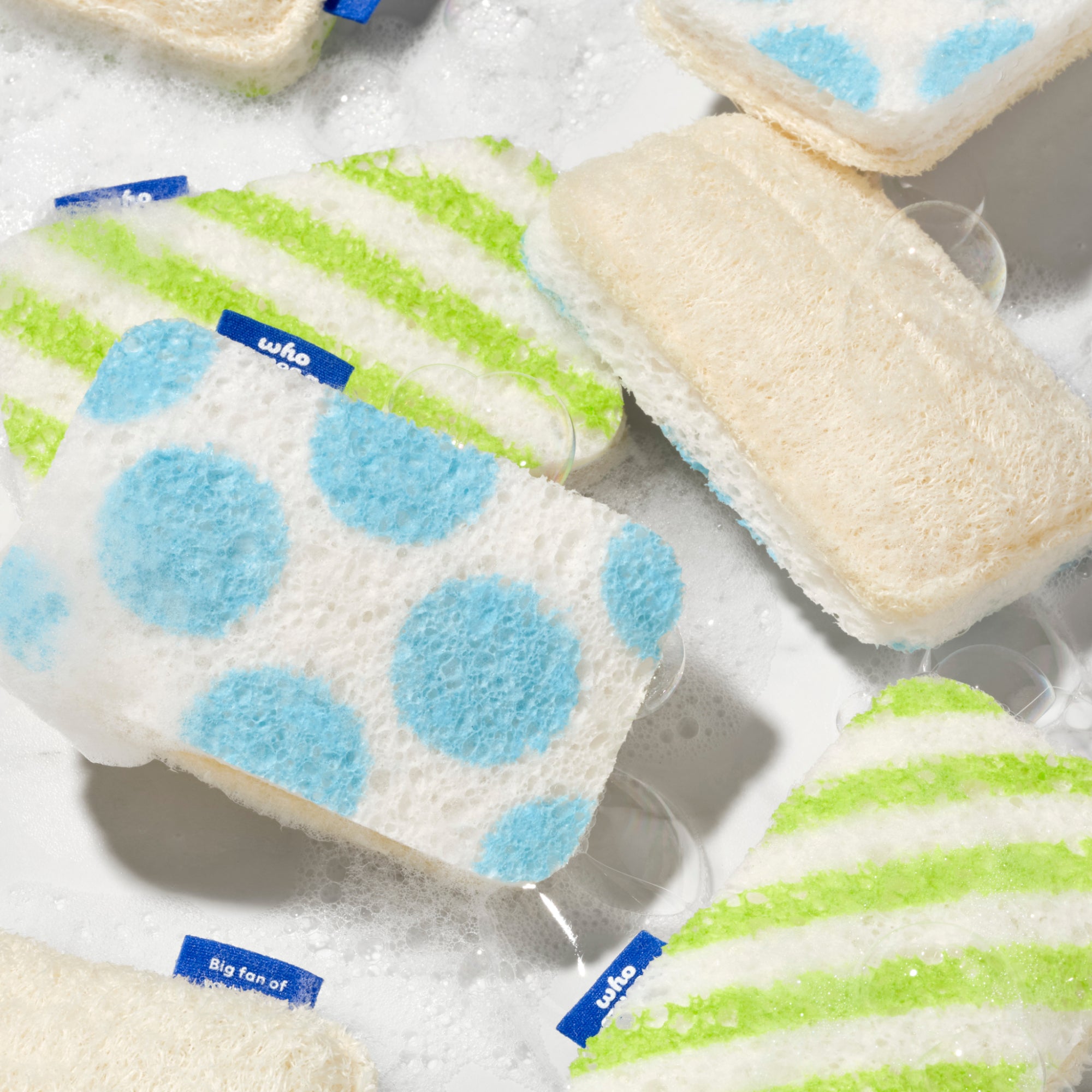 Multiple Who Gives A Crap scourer sponges floating in soapy water, showing the blue polka dot, green stripe and natural loofah designs
