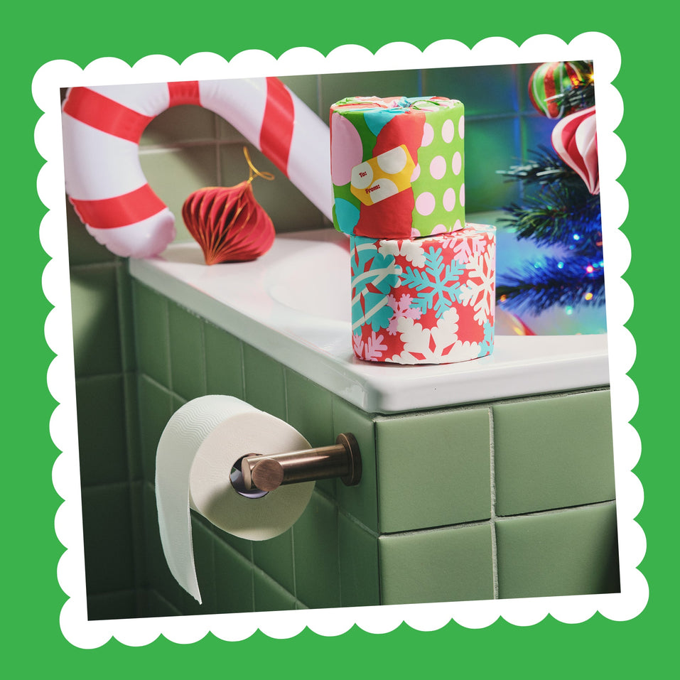 Gift Edition toilet paper rolls in colourful wrappers stacked on a green-tiled bathroom ledge next to a candy cane and a toilet roll holder.