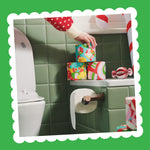 A hand places a wrapped Gift Edition roll on a stack of festive rolls on a bathroom shelf above a toilet roll holder.