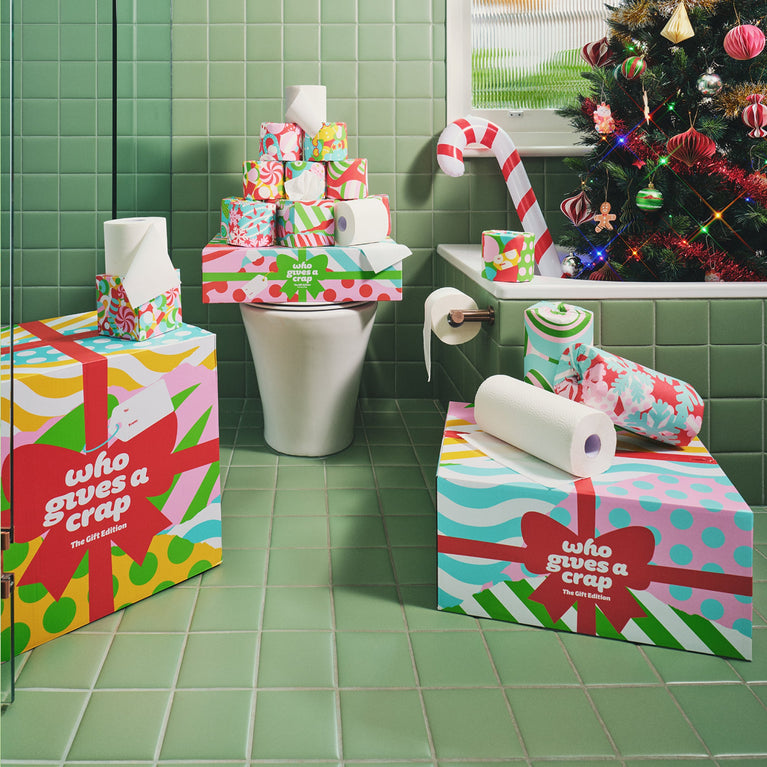 A festive green-tiled bathroom decked out for the holidays, with Who Gives A Crap The Gift Edition toilet paper rolls and boxes in bright gift-wrap designs stacked on the toilet, bath and floor, beside a candy cane and a decorated Christmas tree.