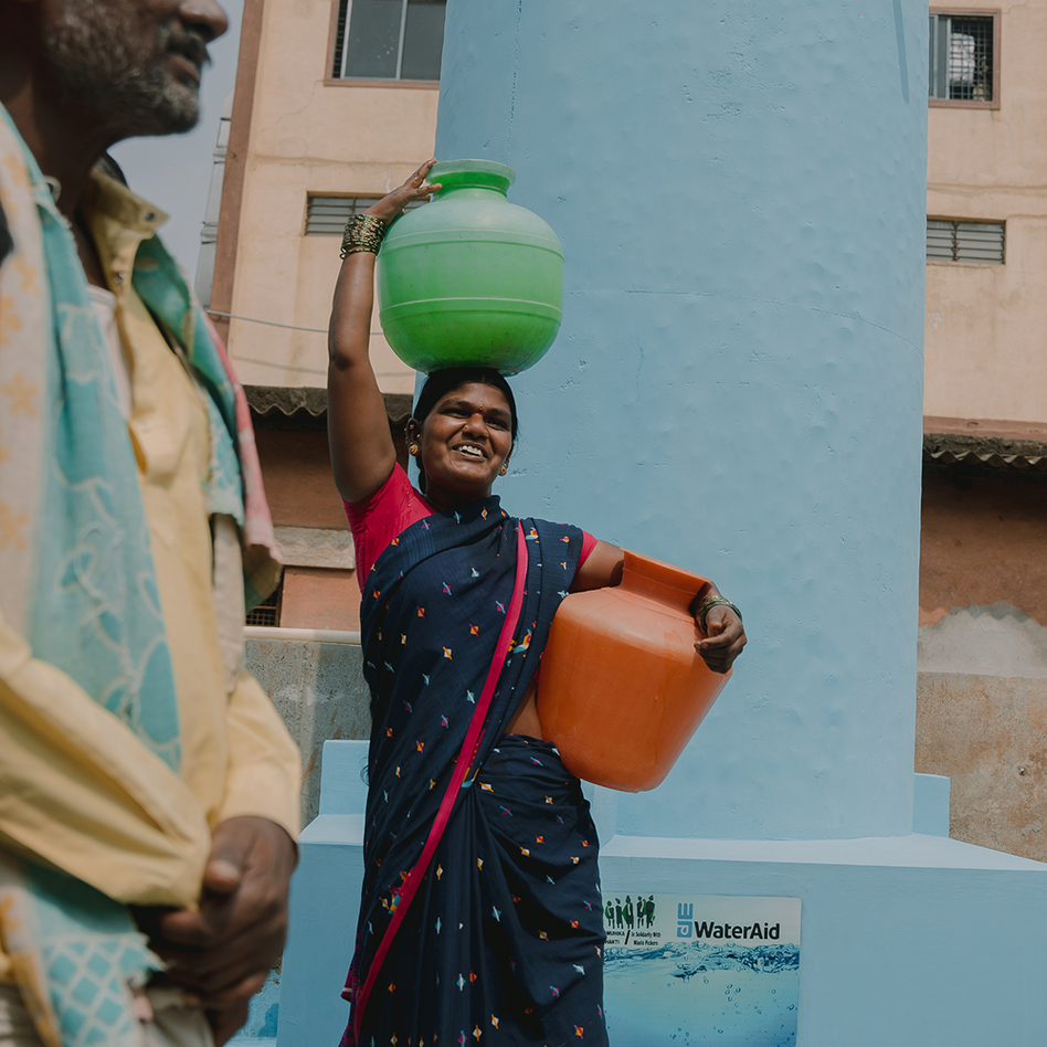 Woman in a blue sari carrying a green water pot on her head and an orange pot at her side, beside a WaterAid water point in India