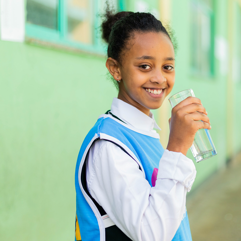 Girl in a blue and white school uniform smiling over her shoulder as she drinks a glass of water, in Ethiopia