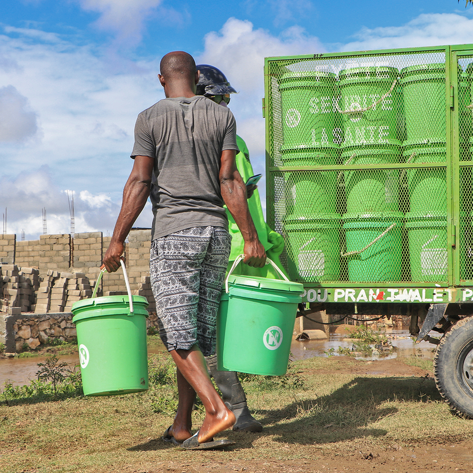 Two workers carrying green toilet containers away from a truck stacked with more of them, in Haiti