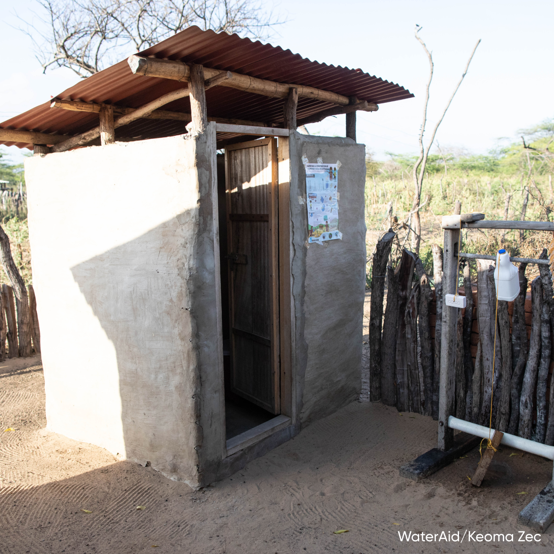 A small toilet outbuilding with some sort of plaster on the outside, a tin roof, and an arid landscape in the background 