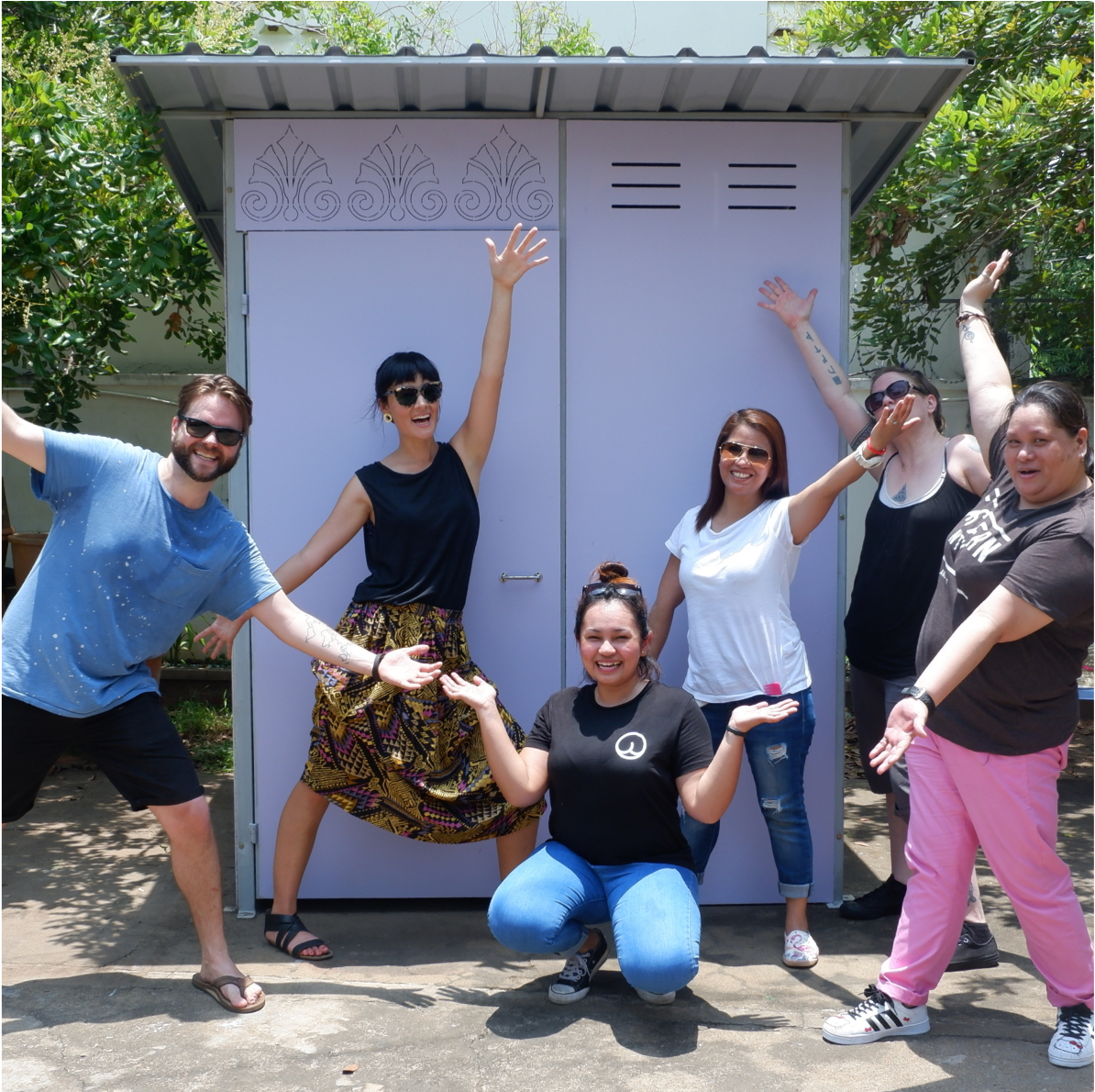 6 members of the Who Gives A Crap team with arms outstretched posing in front of a WaterSHED Ventures portable latrine shelter