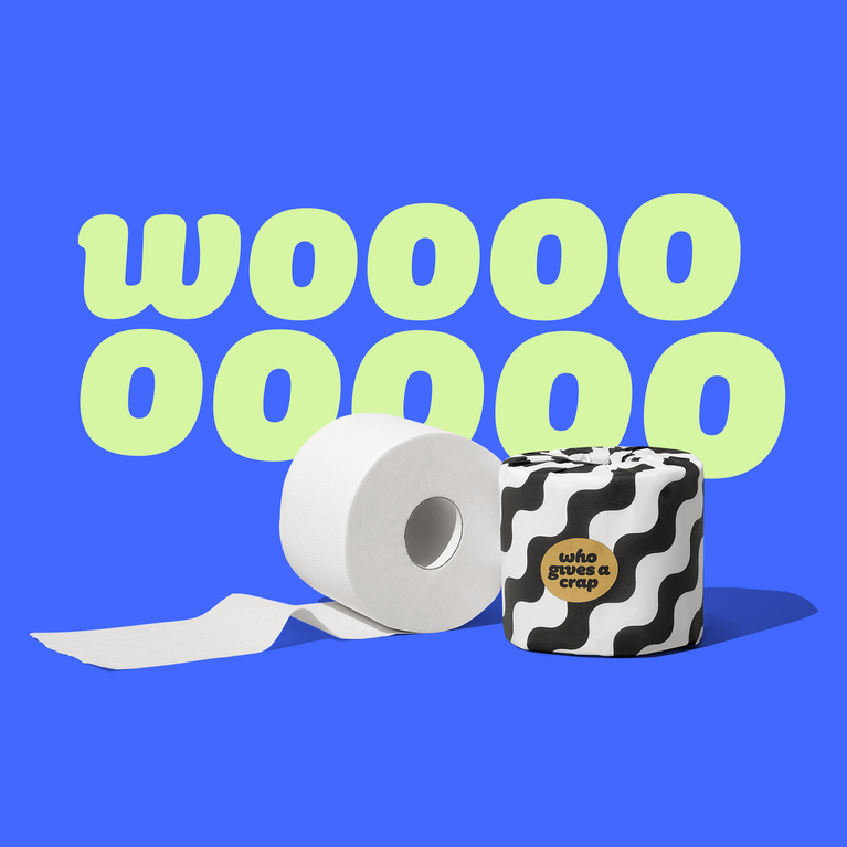 Single roll of open Who Gives a Crap bamboo toilet paper sitting next to a toilet paper roll wrapped in black and white packaging