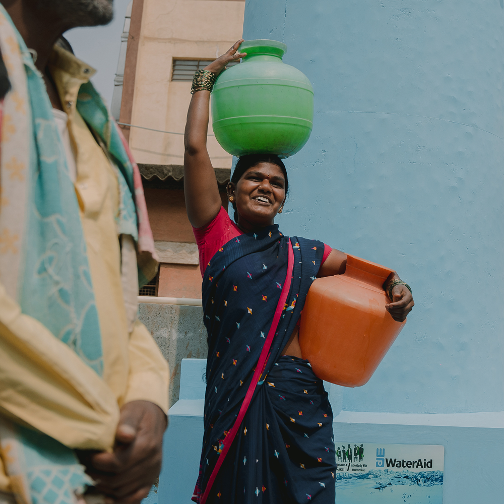 Walking woman carries containers of water on head and hip and smiles