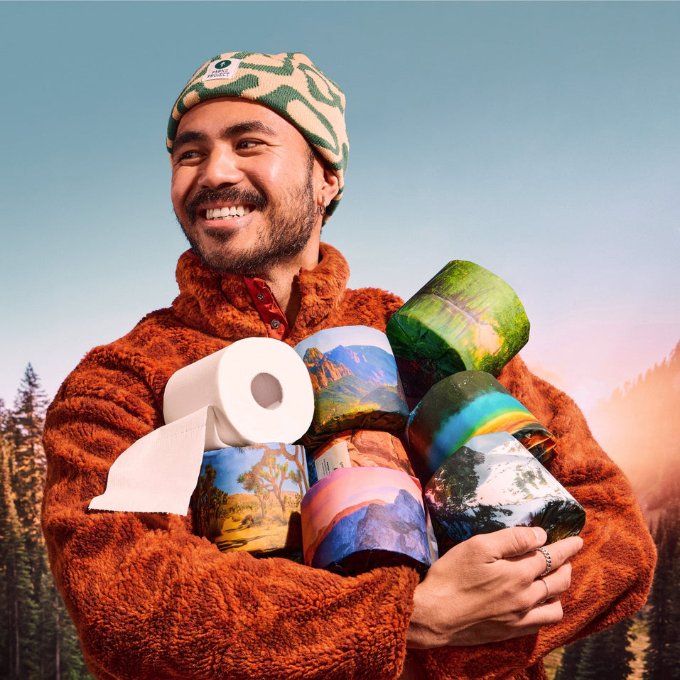 A smiling person in a rust fleece and patterned beanie cradling an armful of park-design rolls plus a white roll, forest and mountains behind.