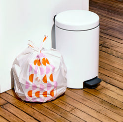 A small white pedal bin on a wooden floor next to a white bin liner printed with a pink and orange semi-circle pattern. 