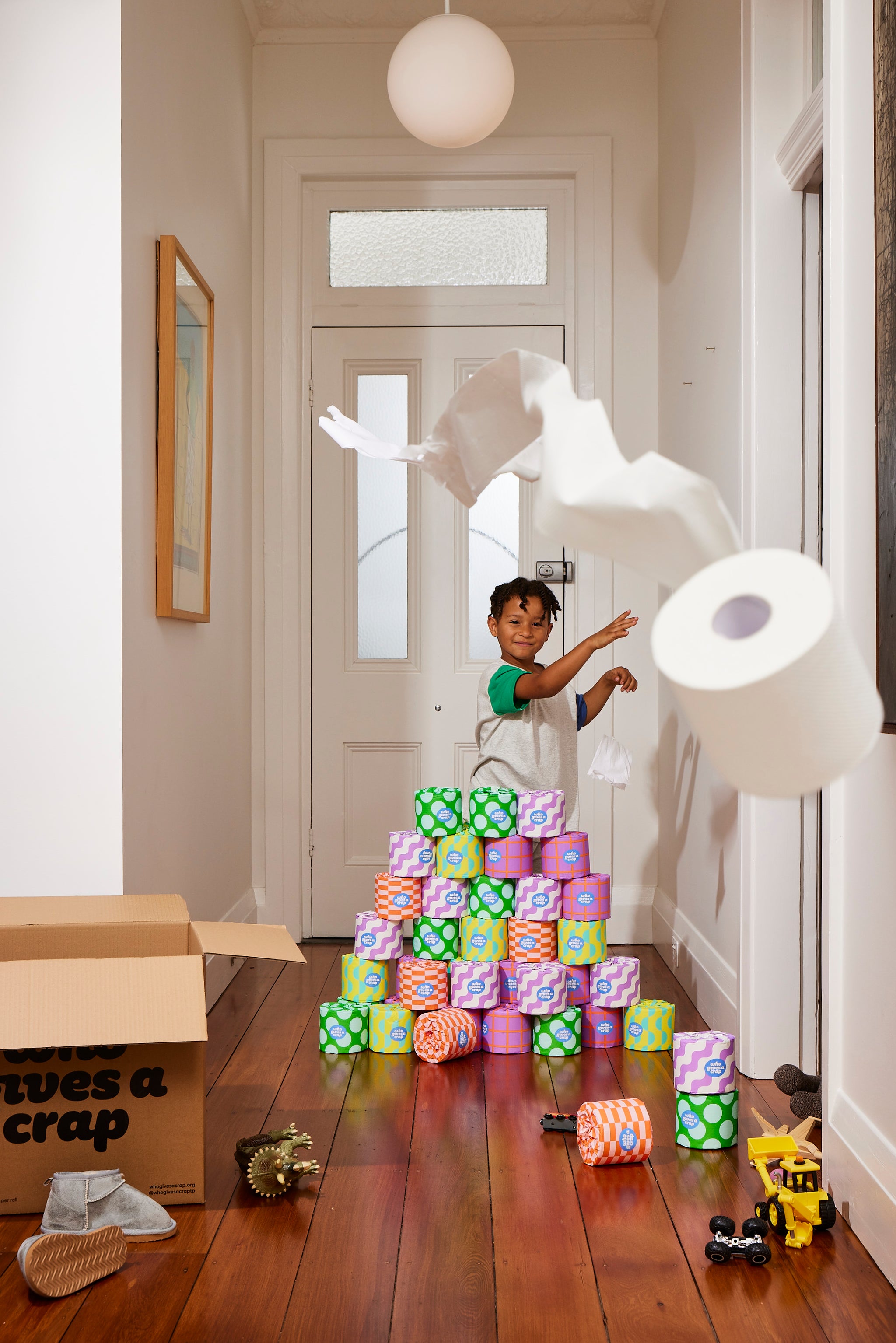 Young smiling boy standing in front of colourful stack of Who Gives A Crap recycled toilet paper playfully throwing toilet paper roll at camera.